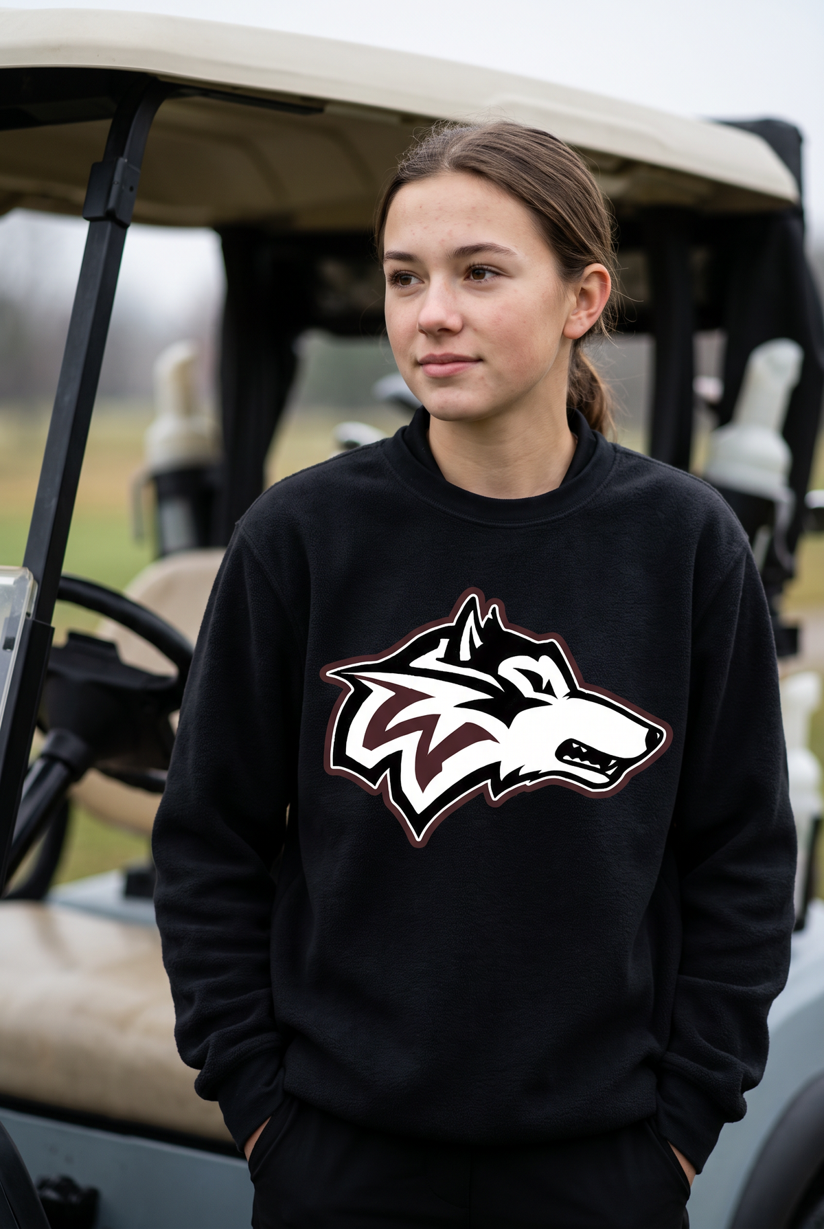 Woodcreek Girls Golf Fleece Crewneck in Black with the Woodcreek wolf head across the chest, worn beside a golf cart